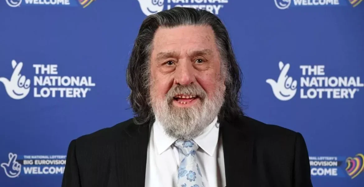 Ricky Tomlinson in a suit and tie posing at a National Lottery event backdrop.