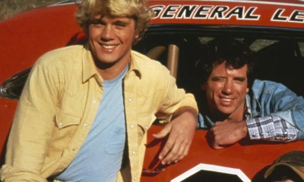 Two men smile near an orange car with the words-tom wopat's net worth