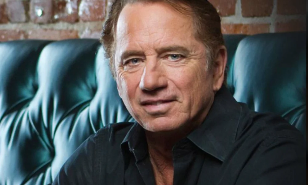 A man with short, light brown hair sits on a dark green leather couch, wearing a black shirt-tom wopat's net worth