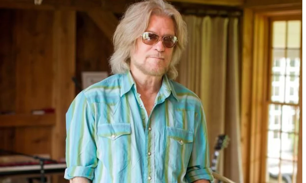 A person with long hair and sunglasses stands in a cozy wooden room-daryl hall net worth