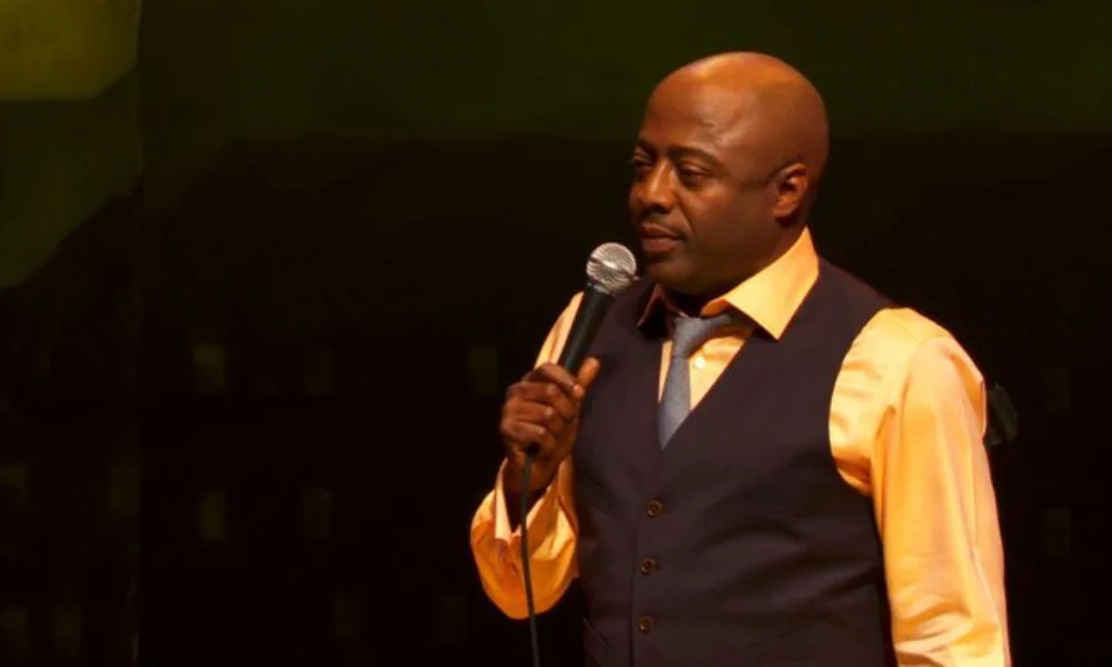 A man in a dark vest and orange shirt holds a microphone on stage-donnell rawlings net worth