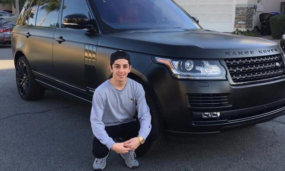 A person smiling and crouching next to a black Range Rover on a sunny street-net worth of faze rug