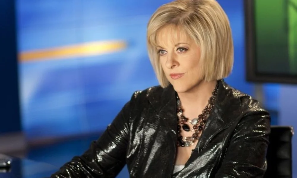 A person with short blonde hair, wearing a shiny black jacket and a statement necklace