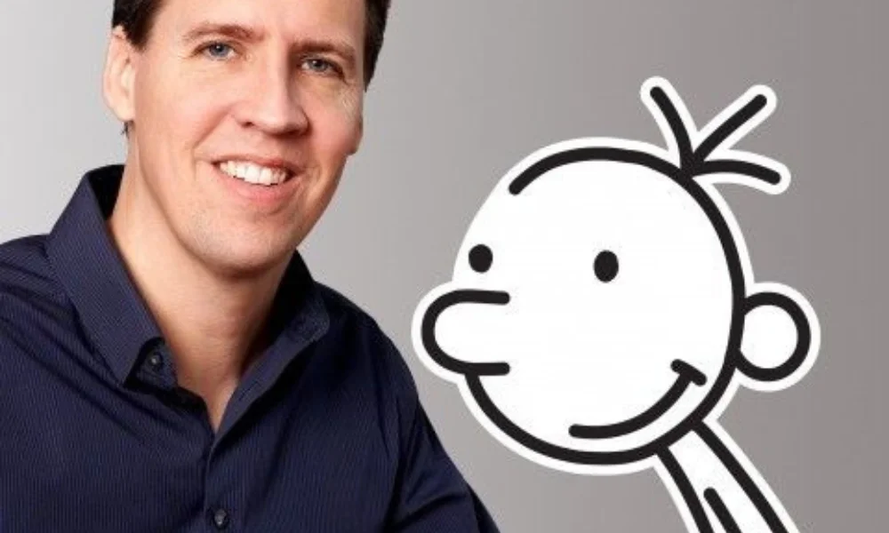 A smiling man in a dark shirt is next to a black-and-white cartoon