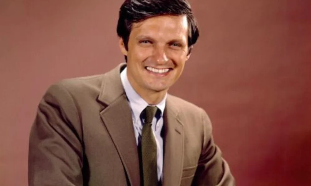 A man in a brown suit and tie smiles warmly-net worth of alan alda