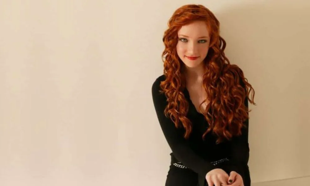 A person with long, wavy red hair wearing a black outfit-scarlett pomers net worth