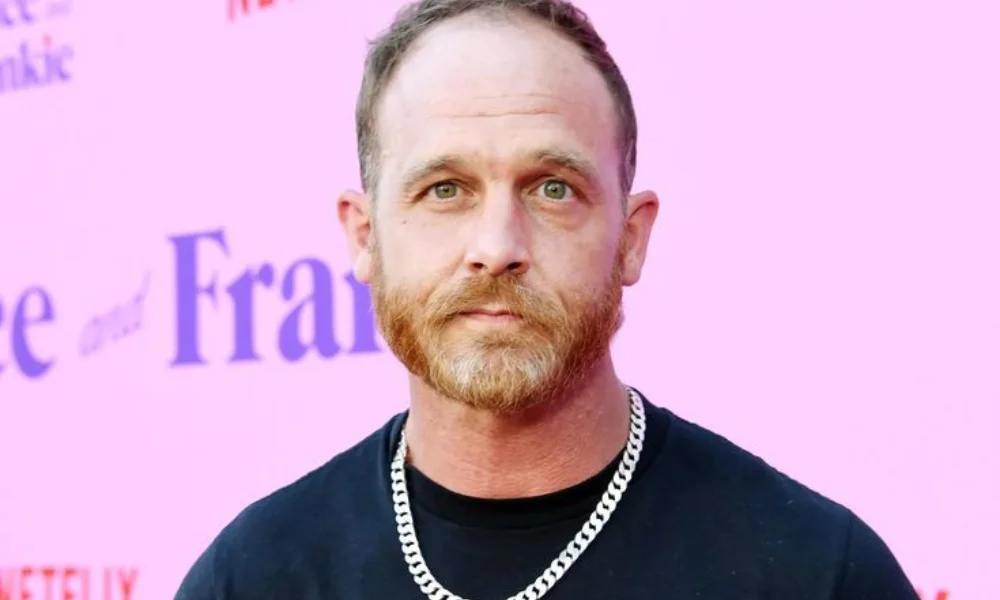 A man with a reddish beard and short hair stands in front of a pink background