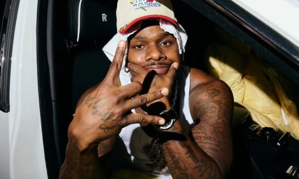 A man in a white tank top and colorful cap sits in a car-dababy's net worth