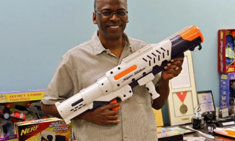 A person smiles while holding a large water gun indoors-lonnie johnson net worth