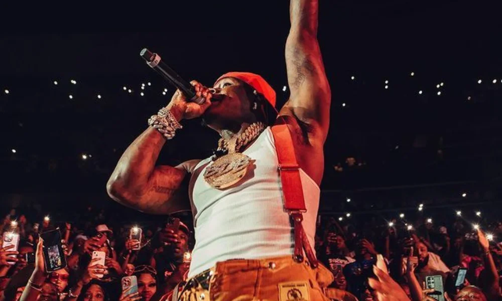 Rapper in a white tank top and orange pants performs energetically on stage, holding a microphone