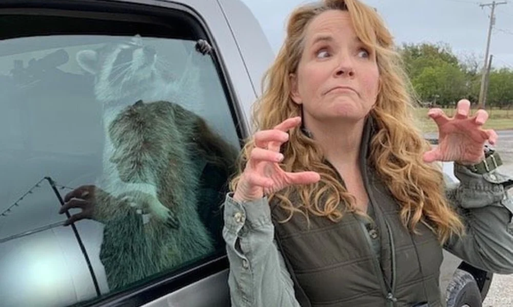A woman with curly blonde hair playfully mimics a raccoon on a car window