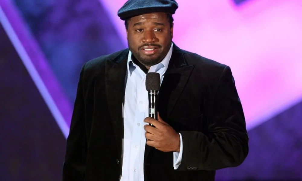 A man in a black jacket and flat cap holds a microphone on stage-corey holcomb net worth