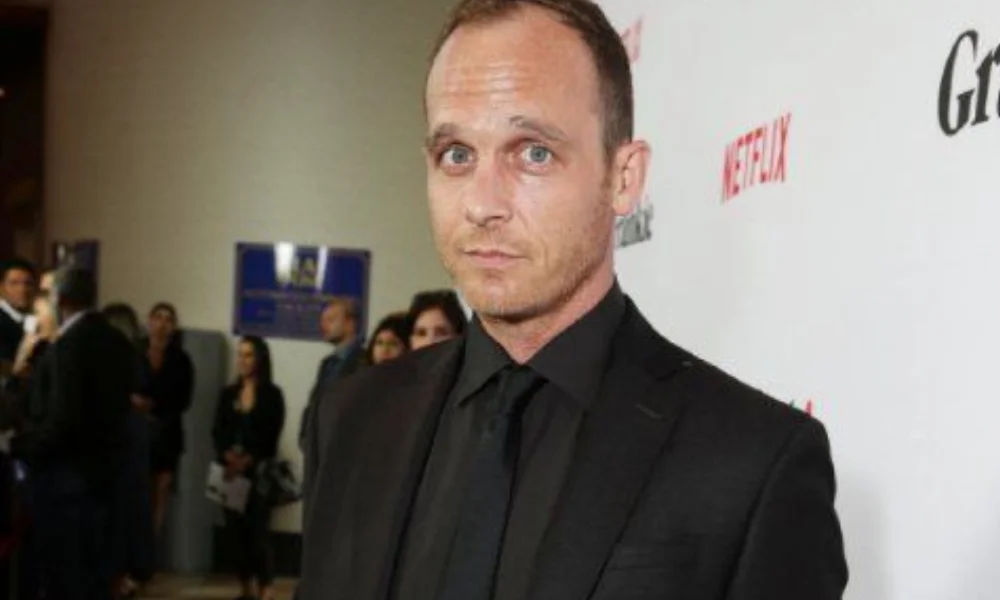 A man in a black suit stands at a formal event, possibly a red carpet-ethan embry's net worth