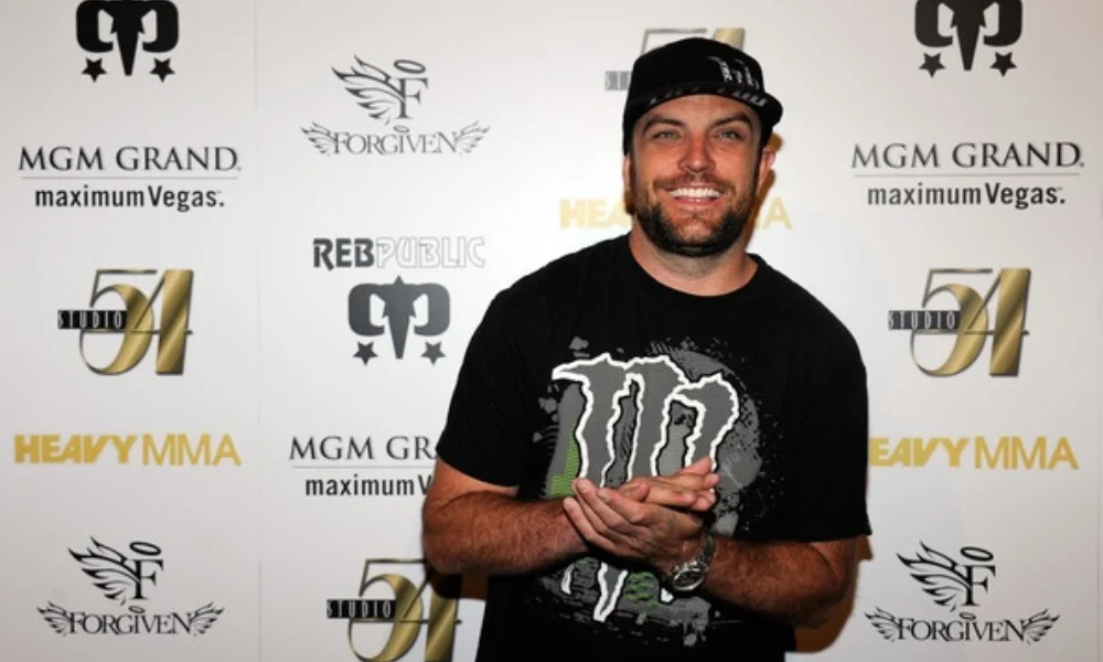 Smiling man in a black cap stands against a backdrop with logos-tj lavin net worth