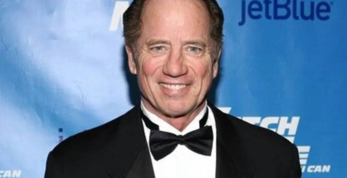 A smiling man in a tuxedo with a black bow tie stands against a blue backdrop