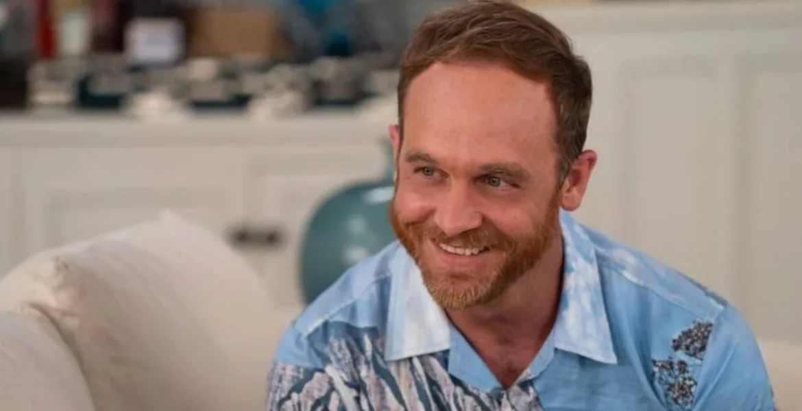 Smiling man with a beard wearing a blue patterned shirt sits on a white sofa