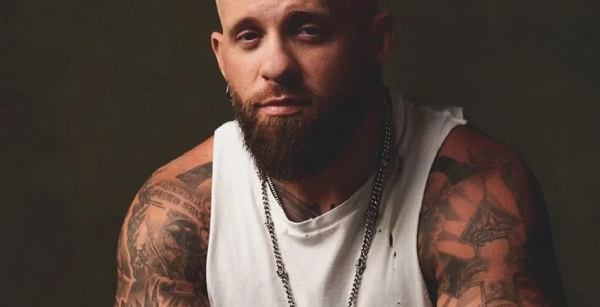 A man with a beard and tattoos sits against a dark background