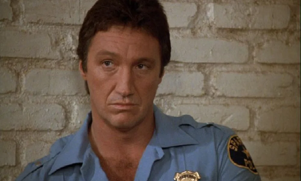 Man in a police uniform leans against a light brick wall
