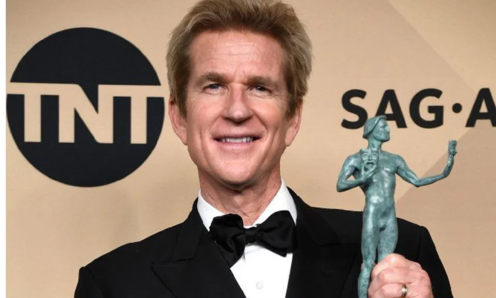 A person in a tuxedo smiles while holding a statuette in a celebratory pose