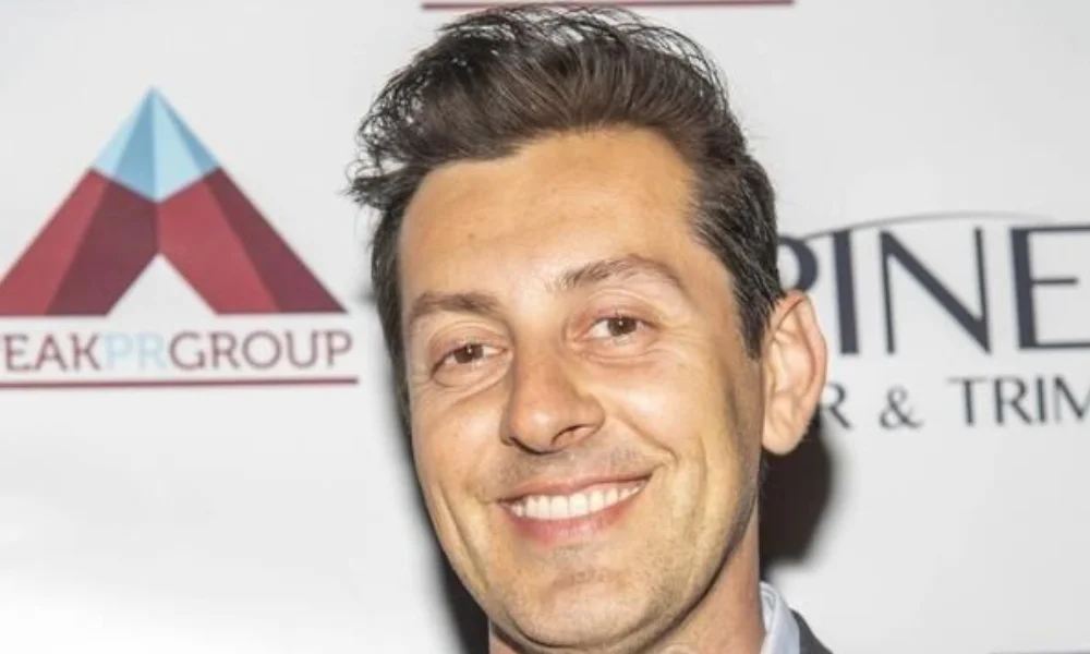 Smiling man with short dark hair in front of a backdrop with logos