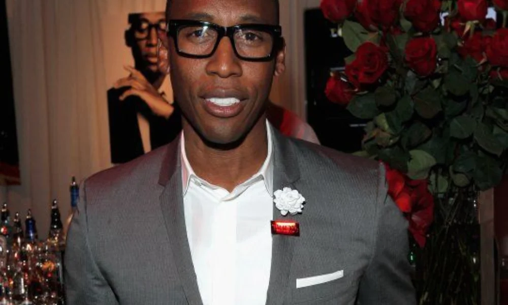 A man in a gray suit with glasses stands smiling