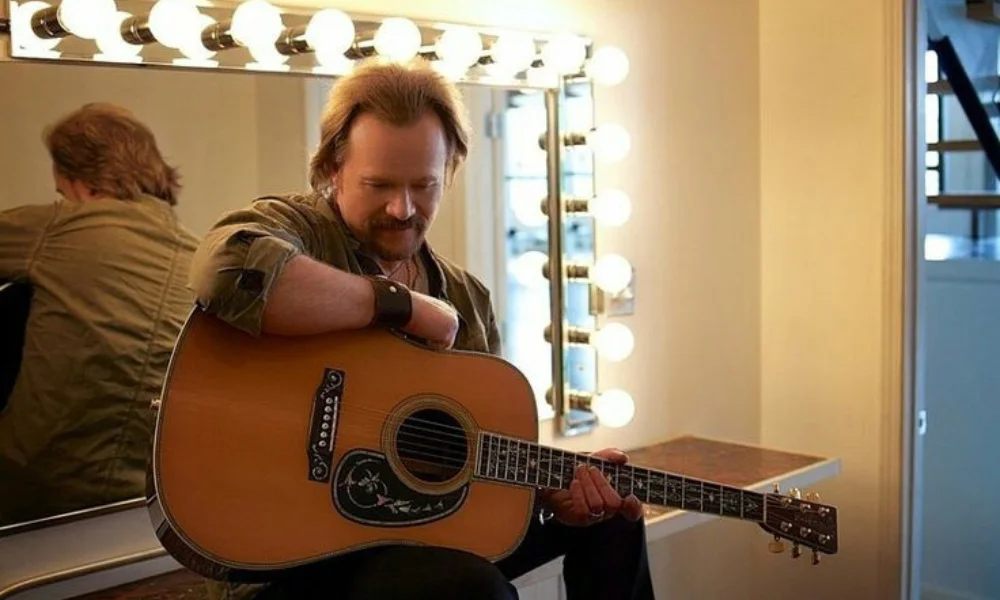 A man with a guitar sits in a warmly lit dressing room-travis tritt net worth