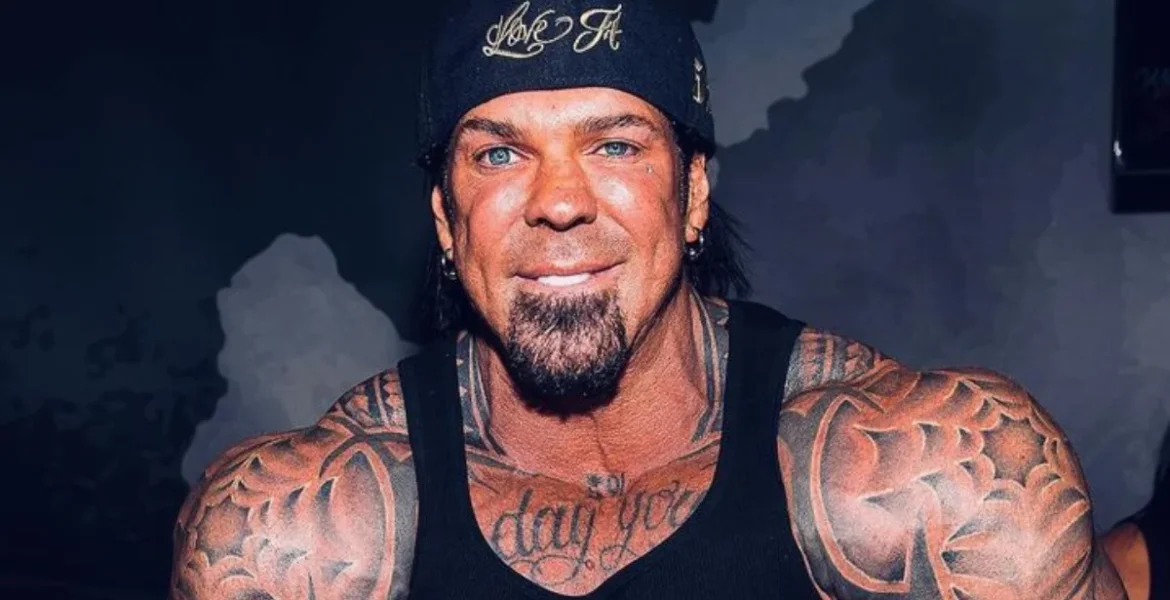 A muscular man with extensive arm tattoos wears a black tank top and cap