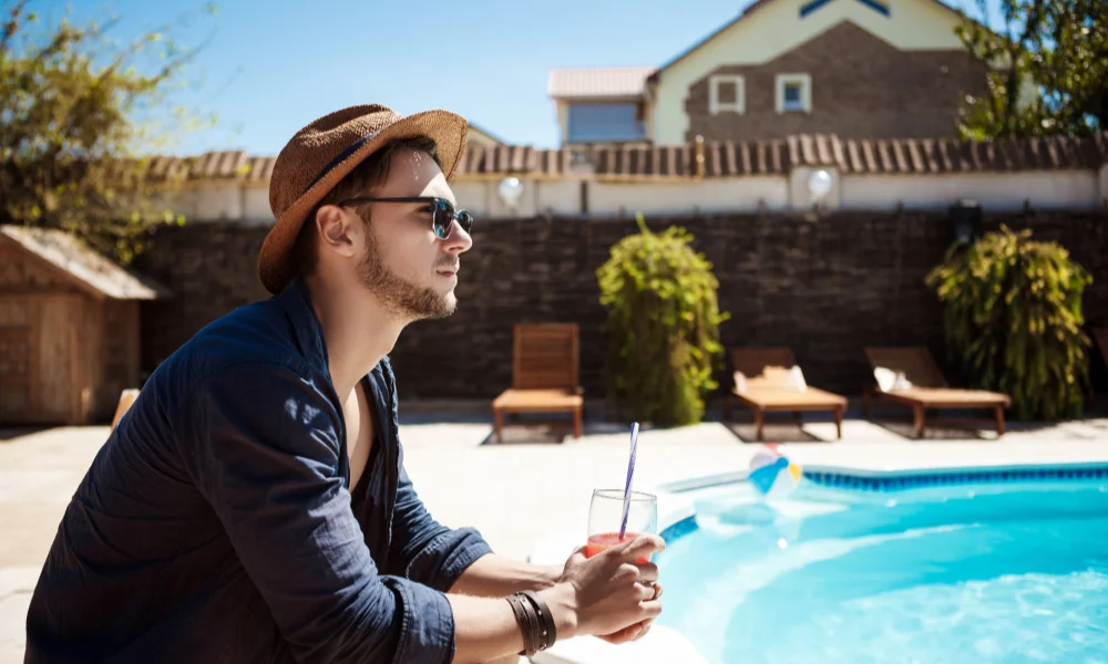 A man in a straw hat and sunglasses relaxes by a bright blue pool-waterfront luxury property